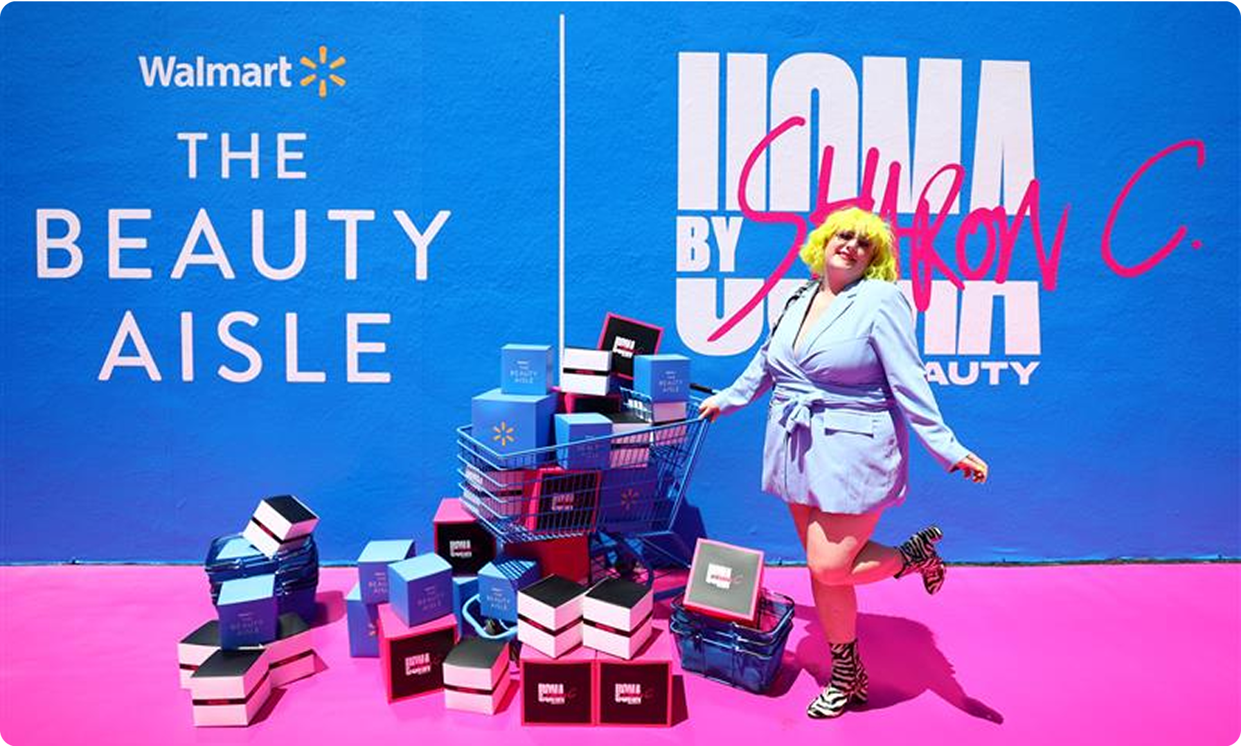 A woman in a stylish blue suit standing with UOMA cosmetics at the Walmart Beauty Aisle event. Standing on a pink carpet.